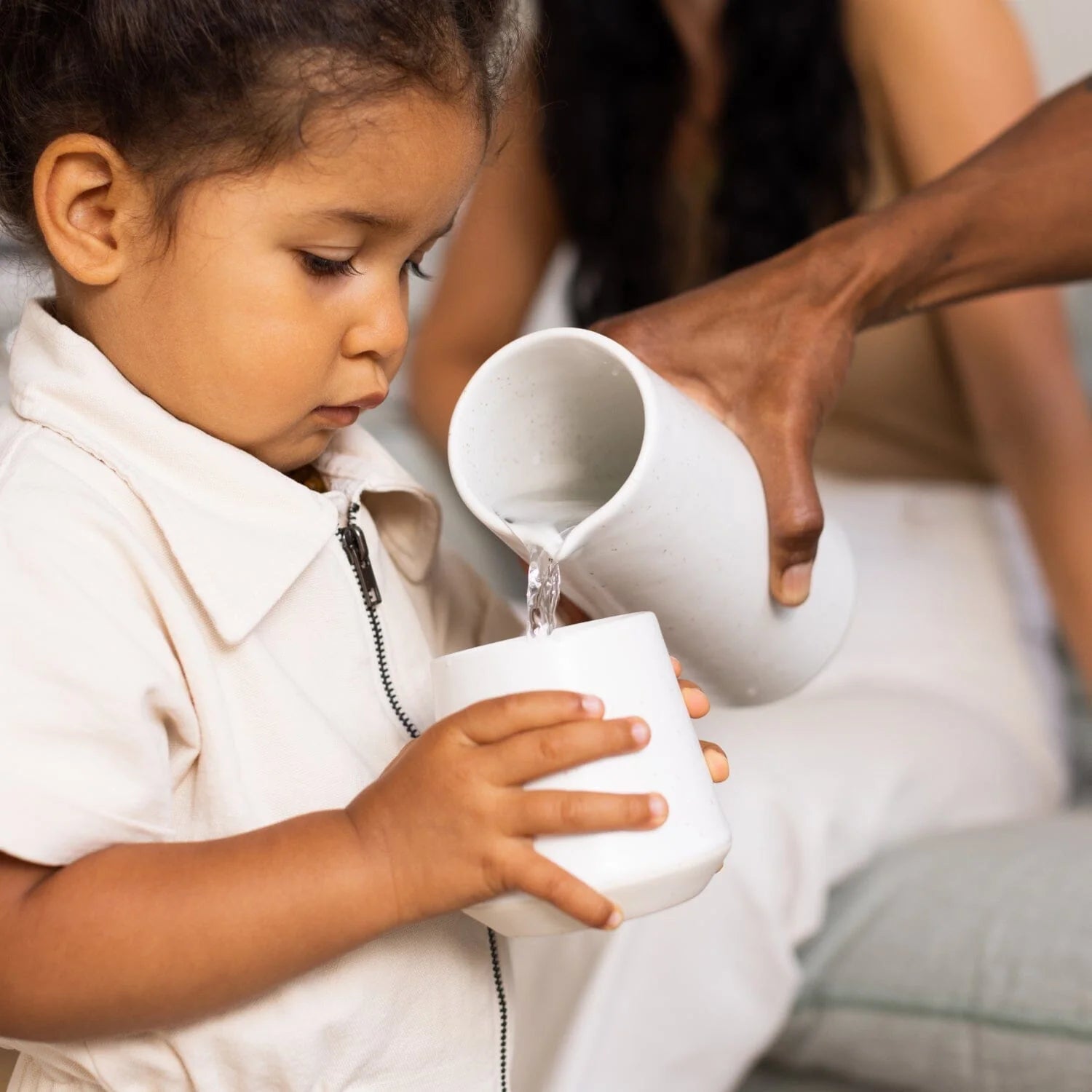 Adult pouring water from one container to another for a child with an adult's hand assisting.