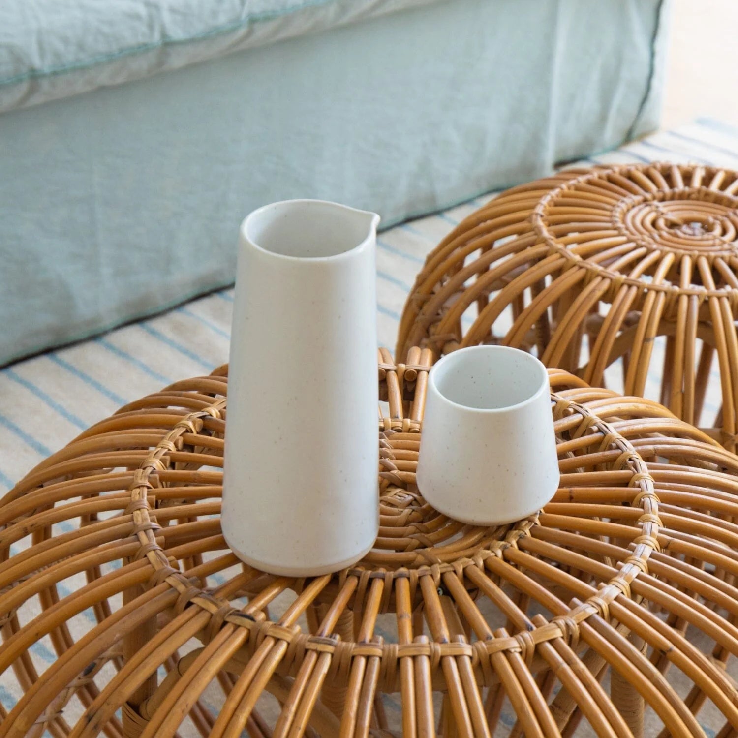 Two white ceramic vases on a woven wooden table with a light blue sofa in the background.