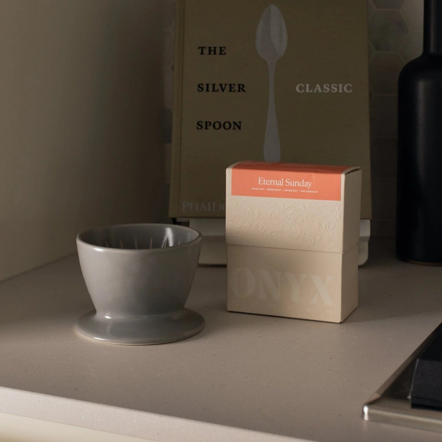 Gray ceramic cup on a surface with a box labeled 'Eternal Sunday' and a book titled 'The Silver Spoon Classic'.